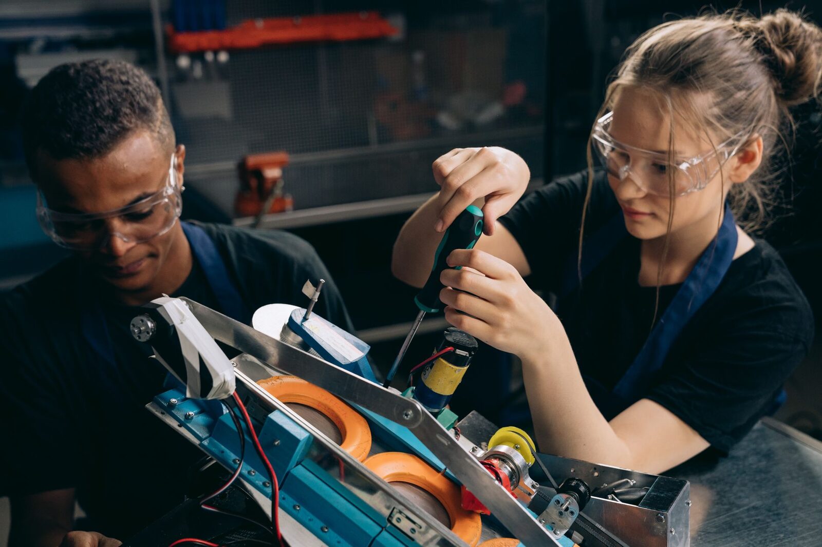Two students in safety glasses assembling a small competition robot with a screwdriver at a lab workbench